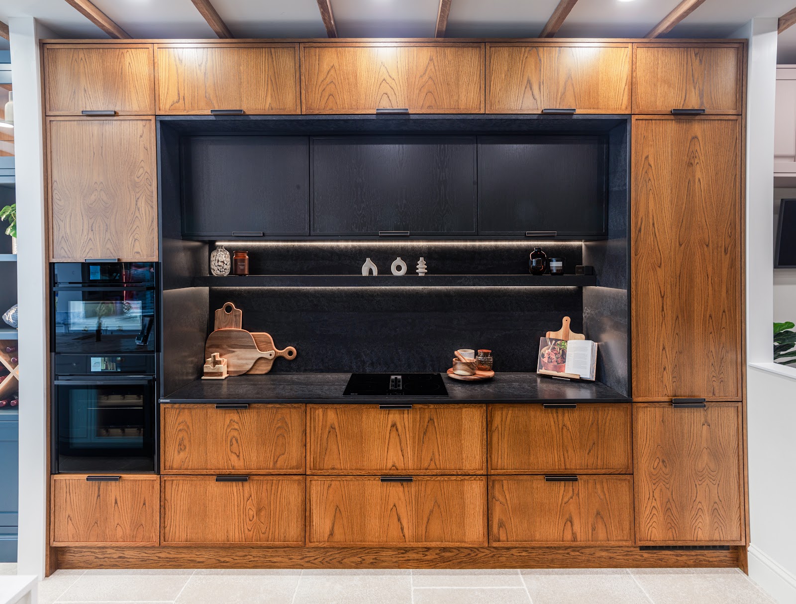 A run from our Premium cabinet tier in oak-veneered timber with black feature cabinetry, integrated ovens and an induction hob set into a dark worktop.