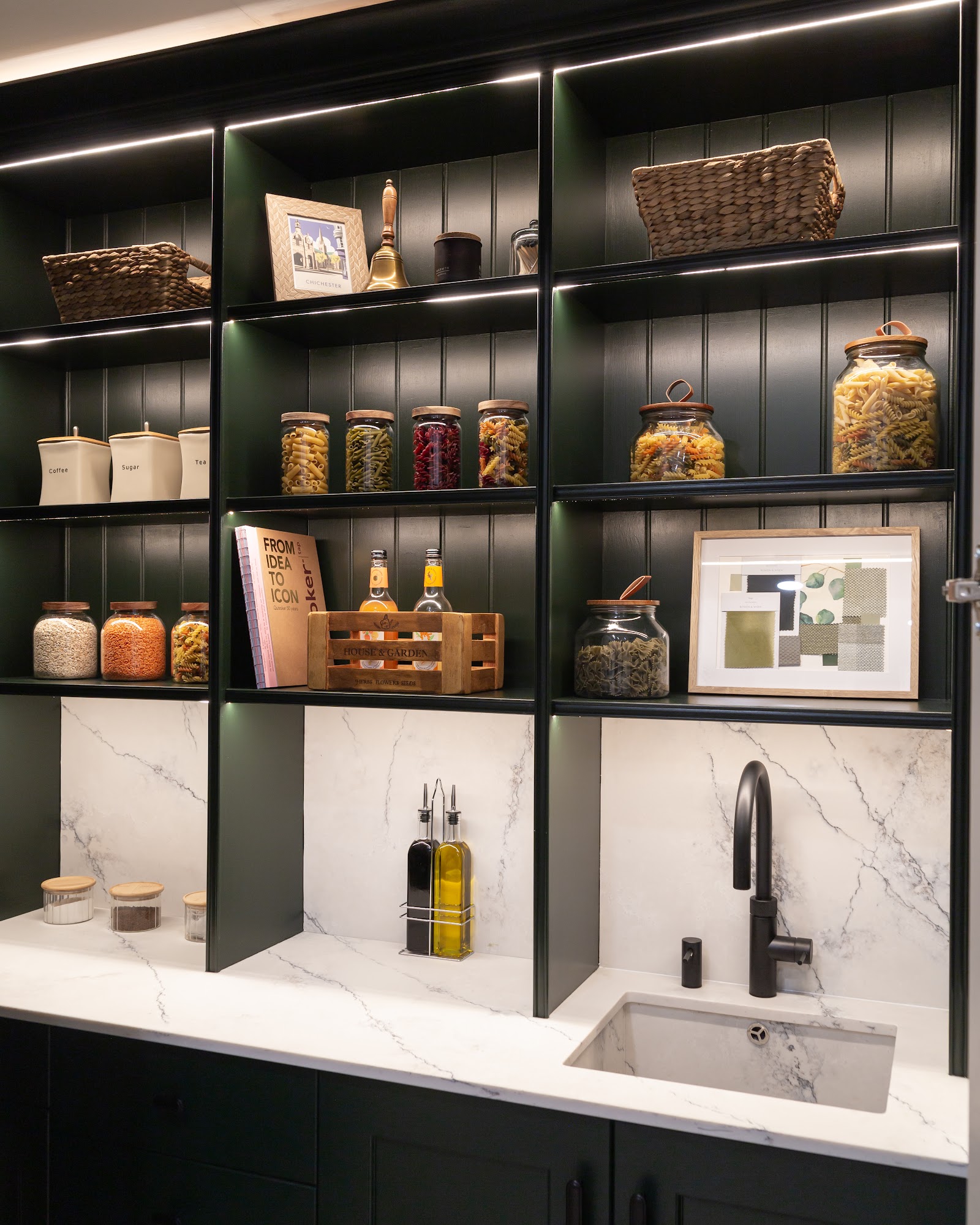 A dark-green larder pantry with lit open shelving, marble-effect splashback, an undermount sink and a matte-black boiling-water tap — storage worked out properly.